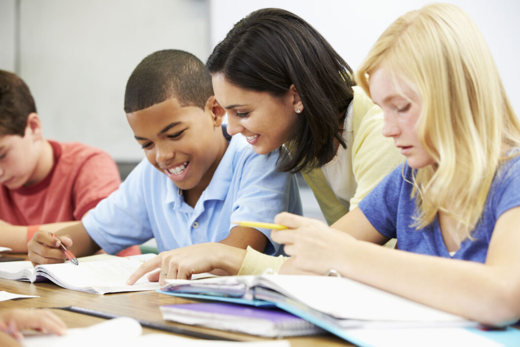 Une enseignante sourit en aidant un élève avec son cahier dans une salle de classe lumineuse.